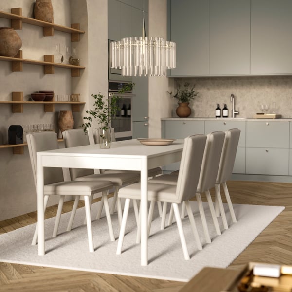 An off-white TONSTAD table with six chairs is on a rug and below a glass chandelier, by an open kitchen with modern fronts.