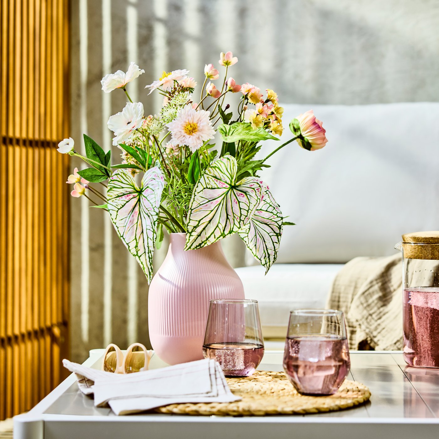 ピンク色の花瓶に生けられた花と葉物がコーヒーテーブルに置かれ、隣にピンク色の飲み物が入ったグラスが2つ並ぶ、明るく居心地の良いリビング。