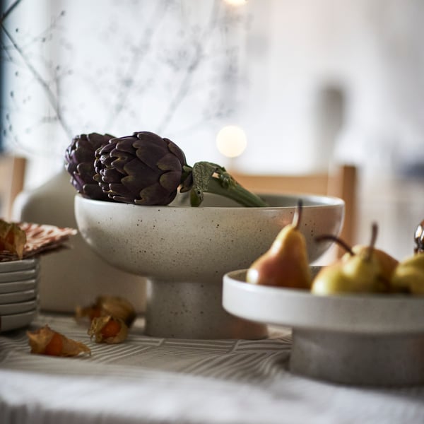 A light beige HÖSTAGILLE serving bowl holds artichokes on a table, next to a HÖSTAGILLE serving stand with pears in it.