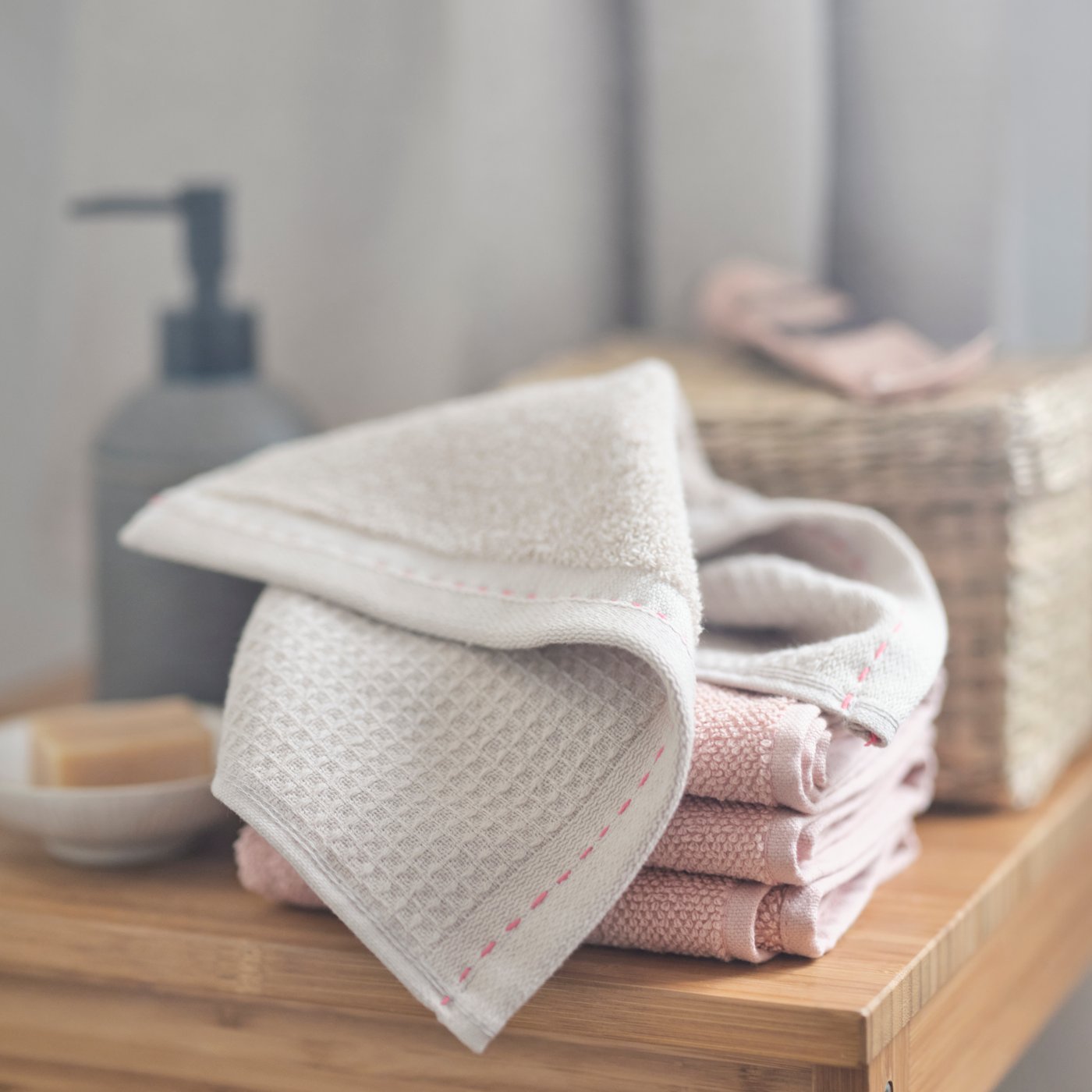 A VÅRDANDE washcloth with hand sewn stitching is folded over a stack of pink towels on a bathroom wooden stool.