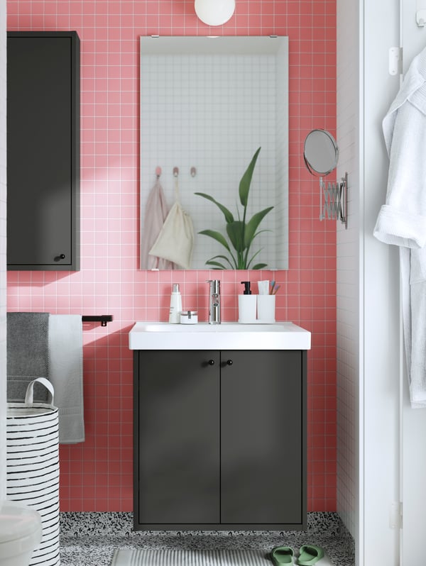 Dans une salle de bains au carrelage rose et au sol à motifs gris, il y a un meuble lavabo HAVBÄCK gris foncé à portes et un miroir au-dessus.