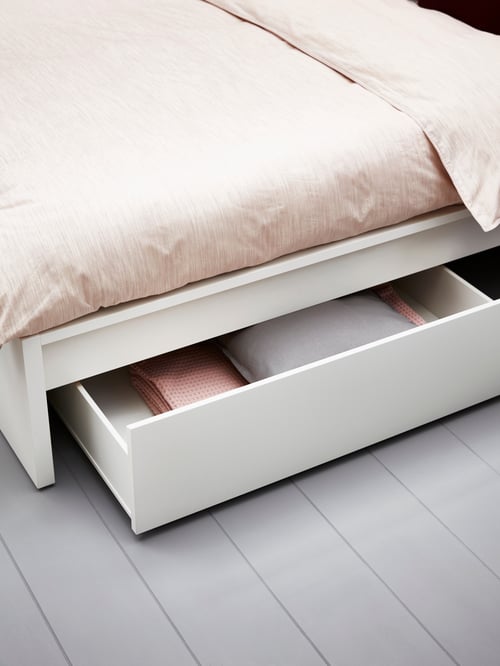 Es ist ein Schlafzimmer mit grauem Fußboden, einem hohen MALM Bettgestell mit 2 Schubkästen in Weiß und rosa Bettwäsche zu sehen. Ein Schubkasten ist ausgezogen und gibt den Blick auf Bettwäsche frei.