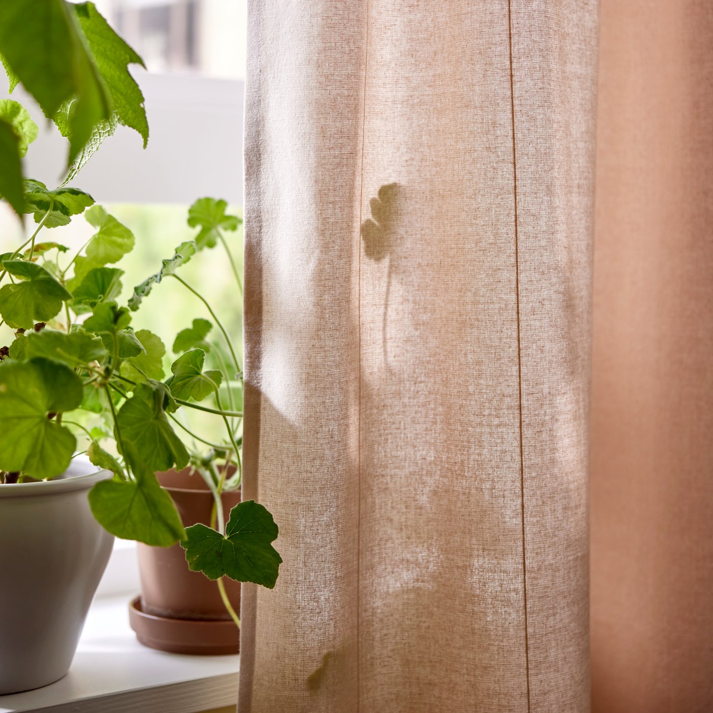 Two plant pots with plants stand on a windowsill with a light pink LENDA curtain hanging at one side of the window.