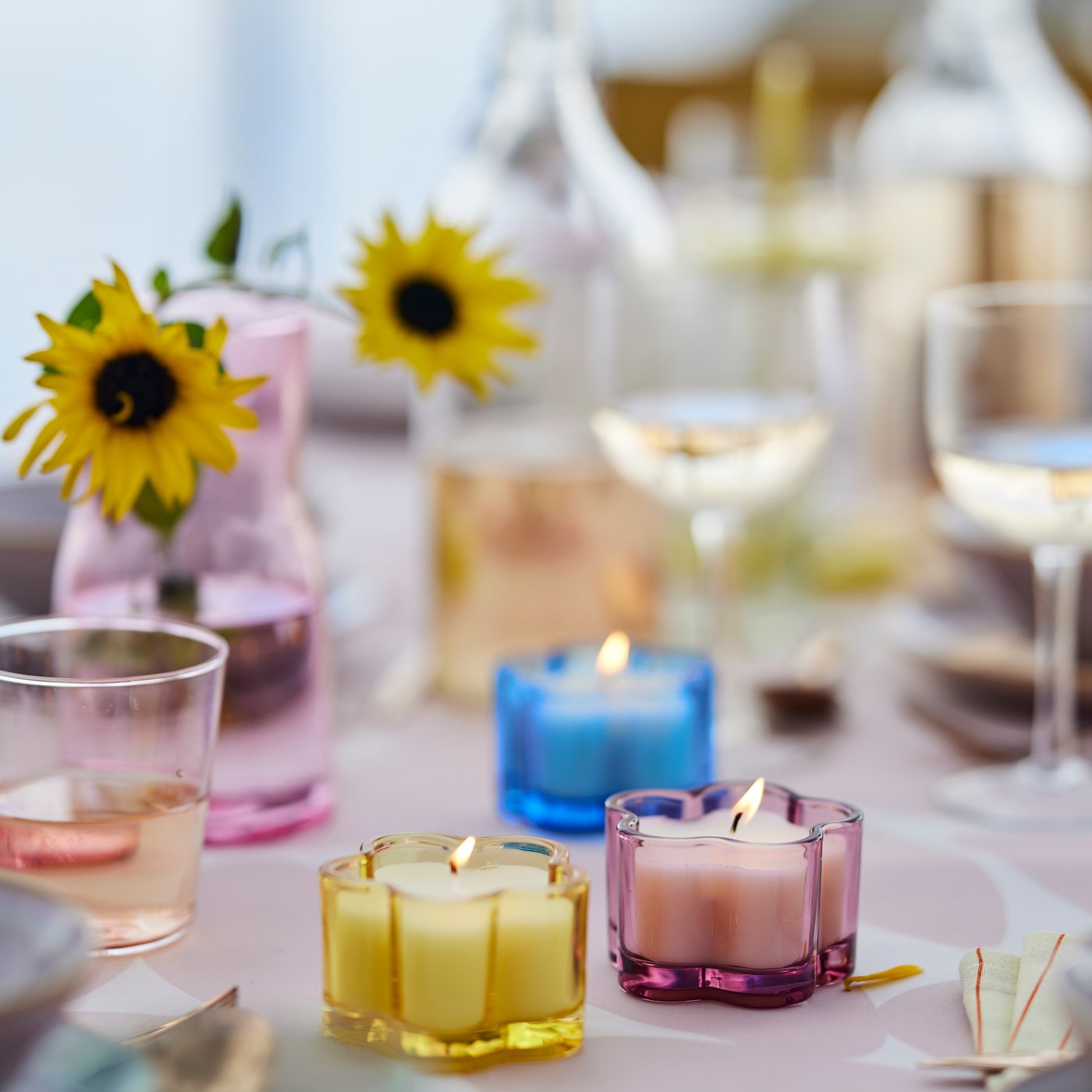 A garden table setting in soft pastels, featuring three MORKULLA floral scented candles in yellow, pink and blue glass.