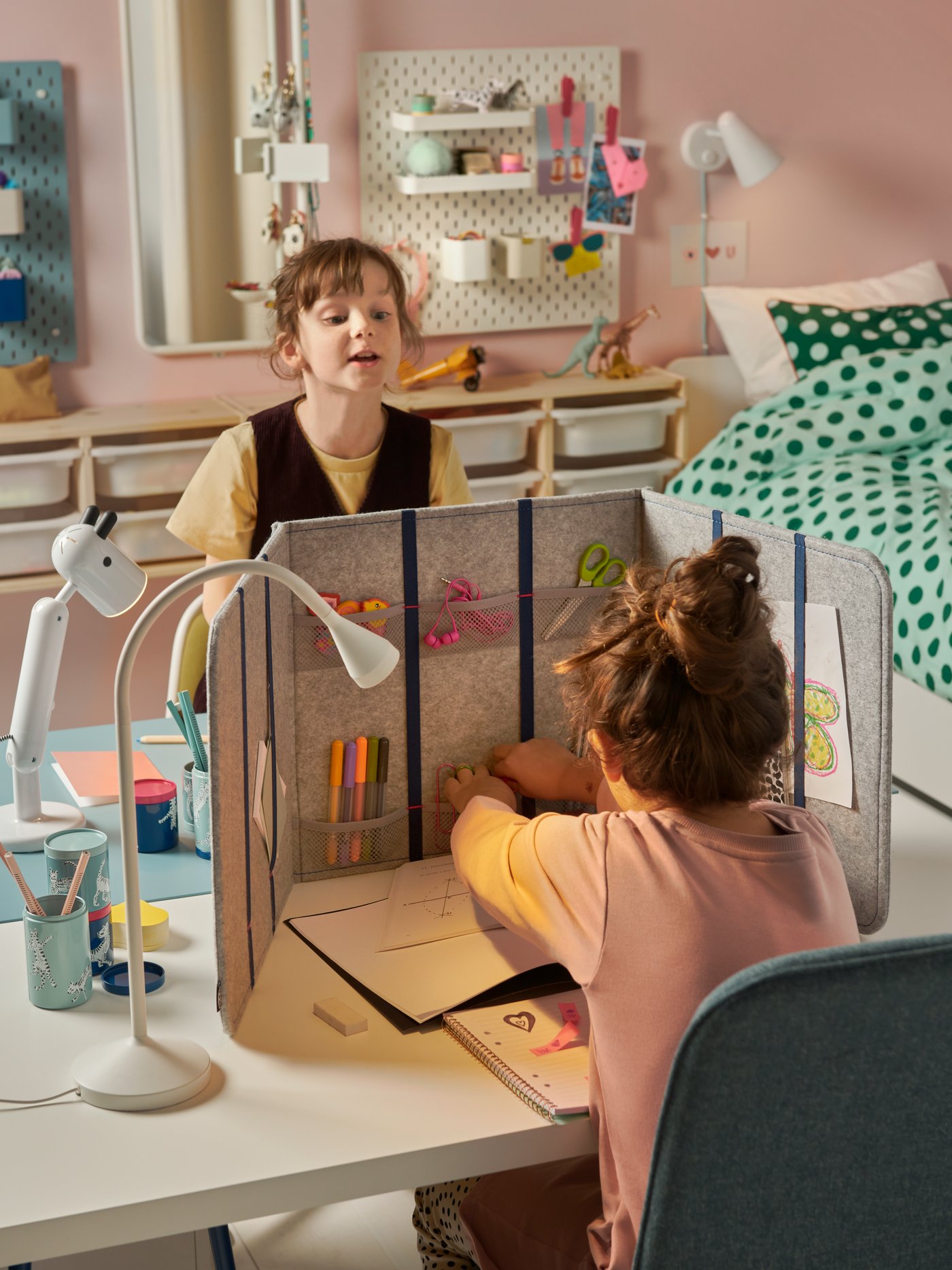 Two girls sit facing each other at opposite sides of two parallel desks. An ÖVNING desk divider stands between the girls.