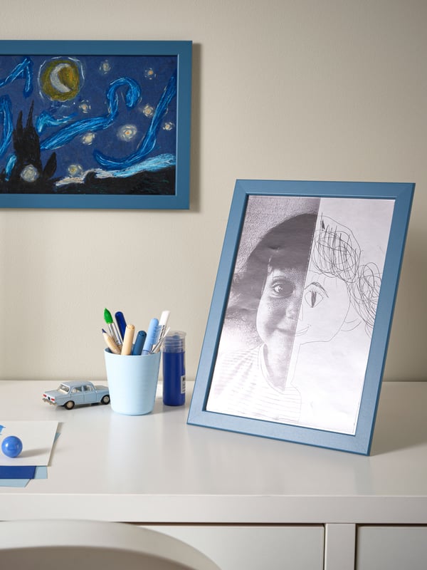 IKEA FISKBO dark grey-blue frame with a drawing on the desk and an IKEA FISKBO dark grey-blue frame hanging on the wall behind the desk.
