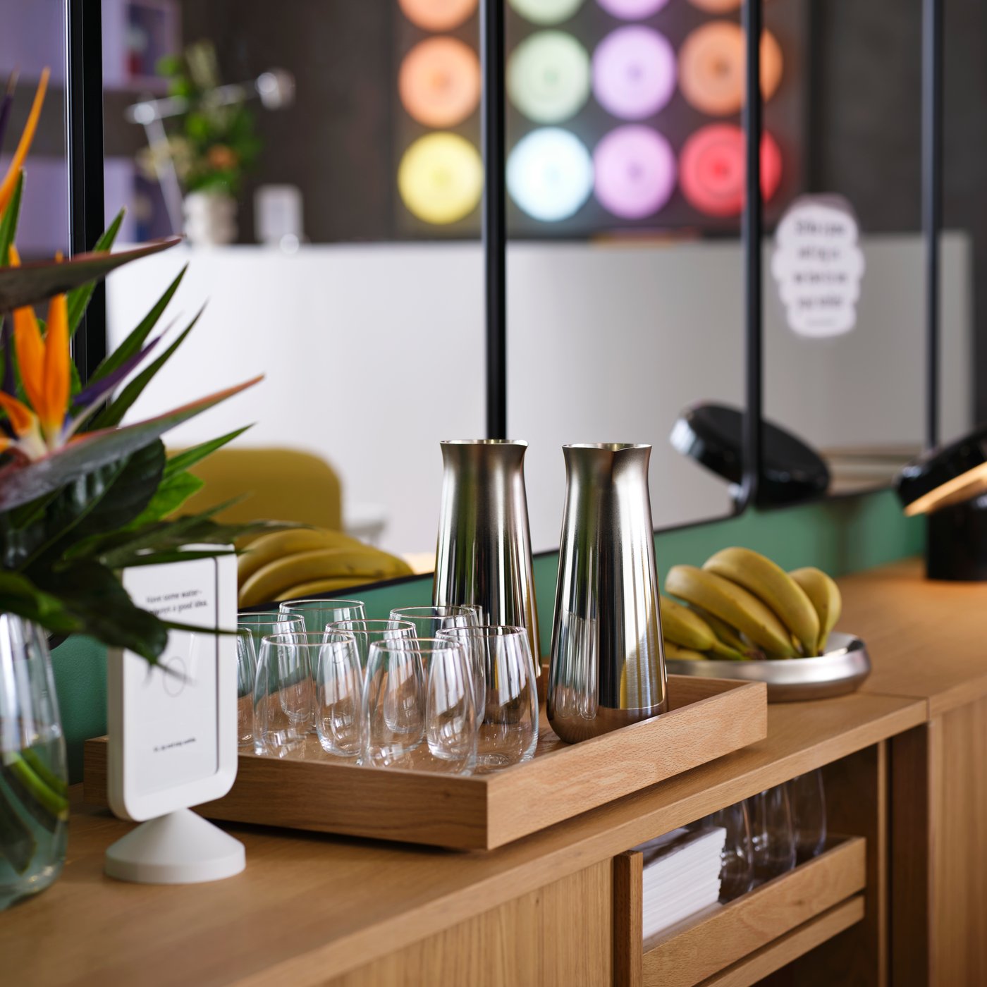 Två eleganta karaffer i rostfritt stål av modellen AFTONHAJ står vid dricksglas och bananer på brickor som en snacksstation i en hotellounge.