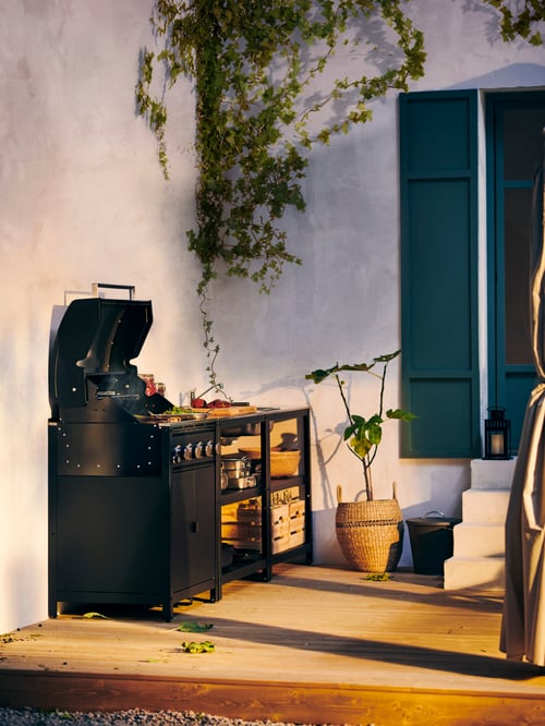 Une cuisine extérieure sur la terrasse d’une maison blanche possède des étagères de rangement et un évier. Le couvercle du barbecue à gaz GRILLSKÄR est ouvert.
