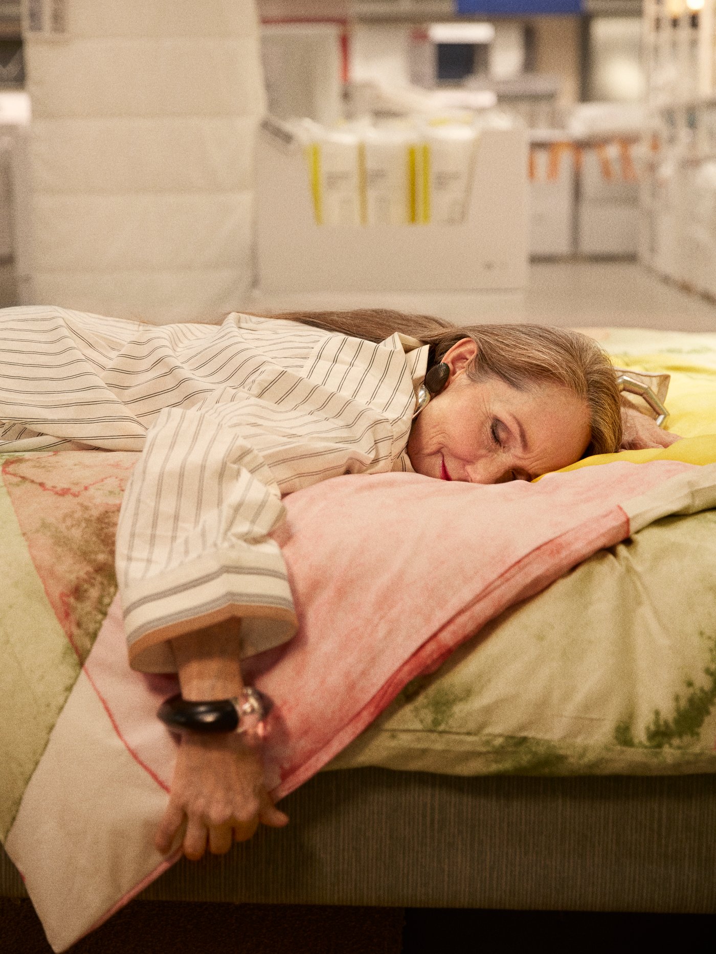 A woman resting with her face down on a comfy bed covered with quilts in an IKEA store's bedroom area.