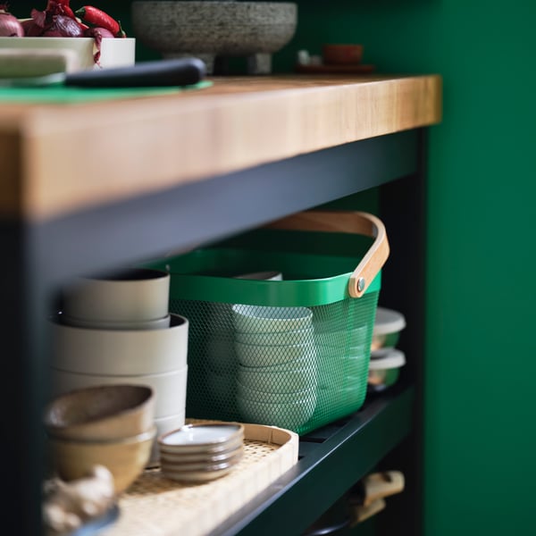 A dark green RISATORP basket with a wooden handle sits on a shelf of a kitchen island next to a tray, plates, and bowls.