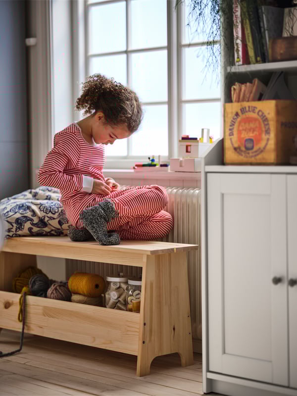 A girl sits on a PERJOHAN pine bench that stands beneath a window. Beside the window stands a white HAUGA high cabinet.