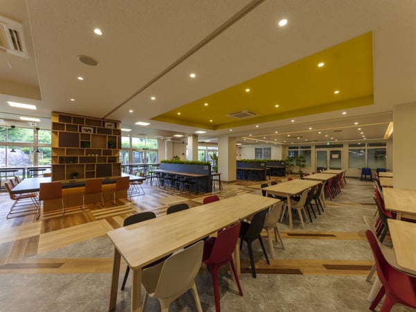 明るい黄色の天井パネルの下で、多くの人がテーブル木製床と大きな窓がある明るい室内で、長テーブルと椅子が整然と並ぶ多目的スペースに座って交流している広々とした室内
