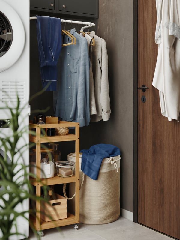 A NATTGIBBA willow laundry basket is standing next to a washing machine and a dryer, below a white rack with hanging shirts.