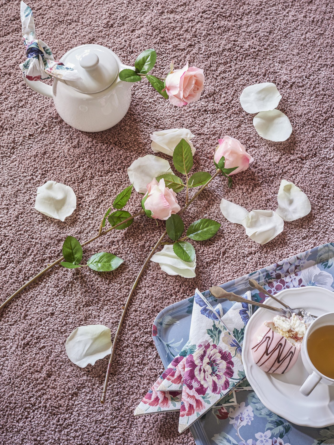 Pink and white flowers and petals lying on the floor next to a tray and teapot on a pink rug.