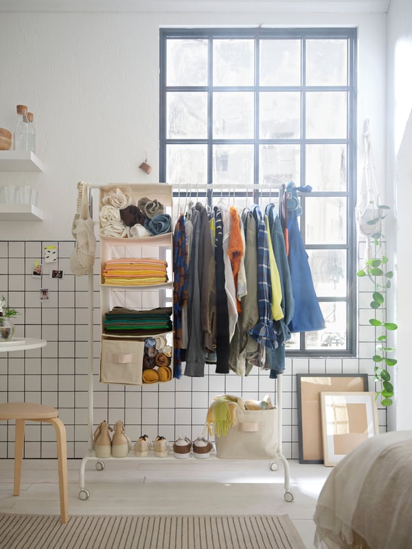 A white RIGGA clothes rack in a white, tiled room, complemented with PURRPINGLA hanging storage and storage baskets.