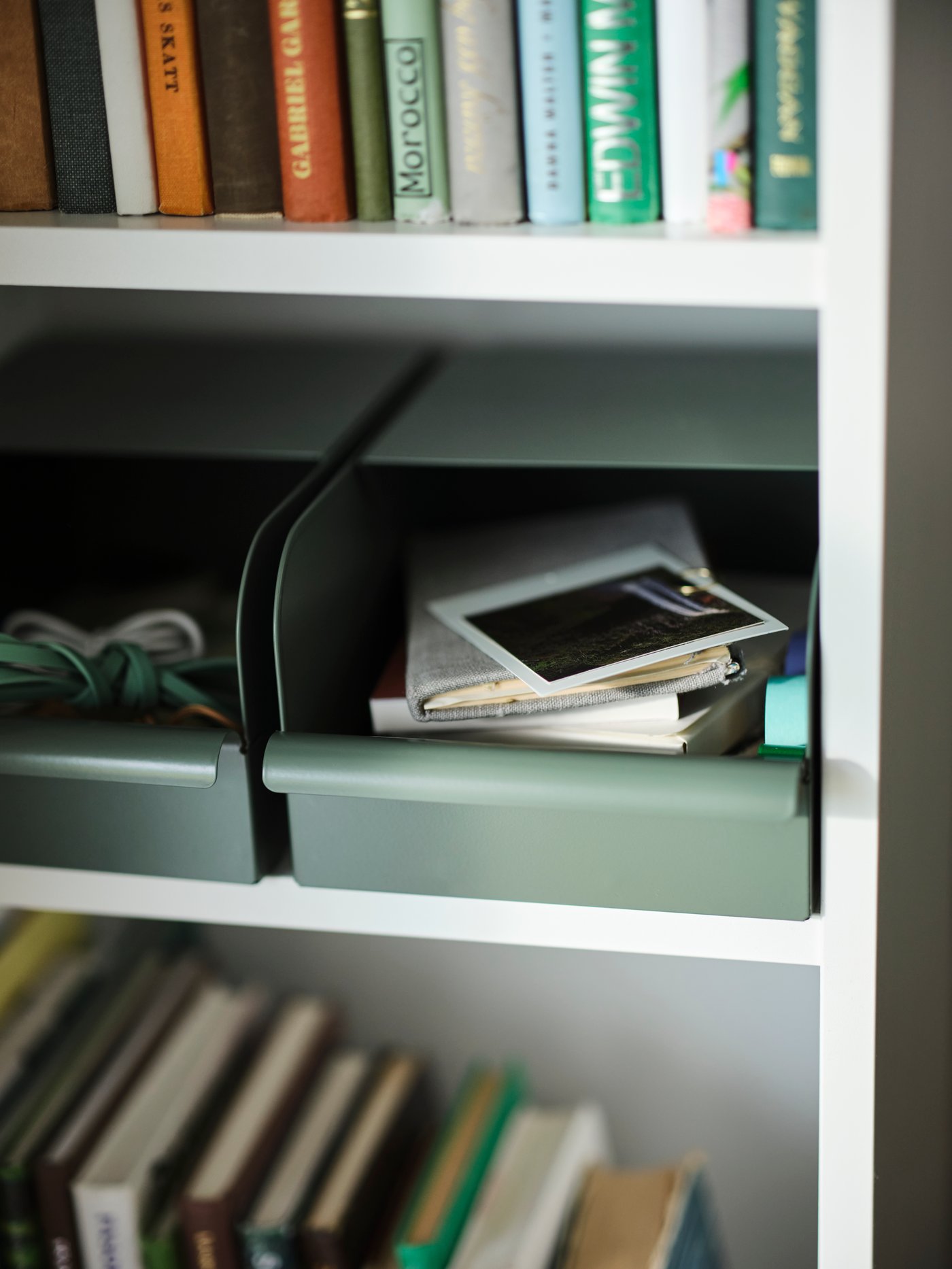 Two grey-green metal REJSA boxes on a white shelf, holding small notebooks, cables and a polaroid photograph.