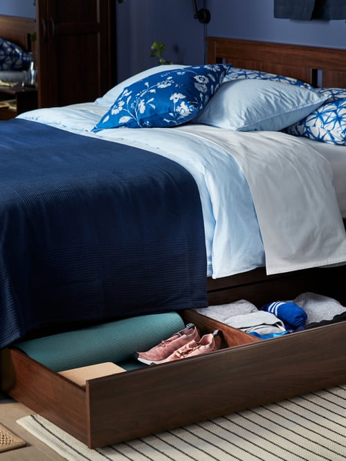 A close-up of a big, open storage drawer with clothes and shoes under a brown SONGESAND bed frame with four storage boxes.