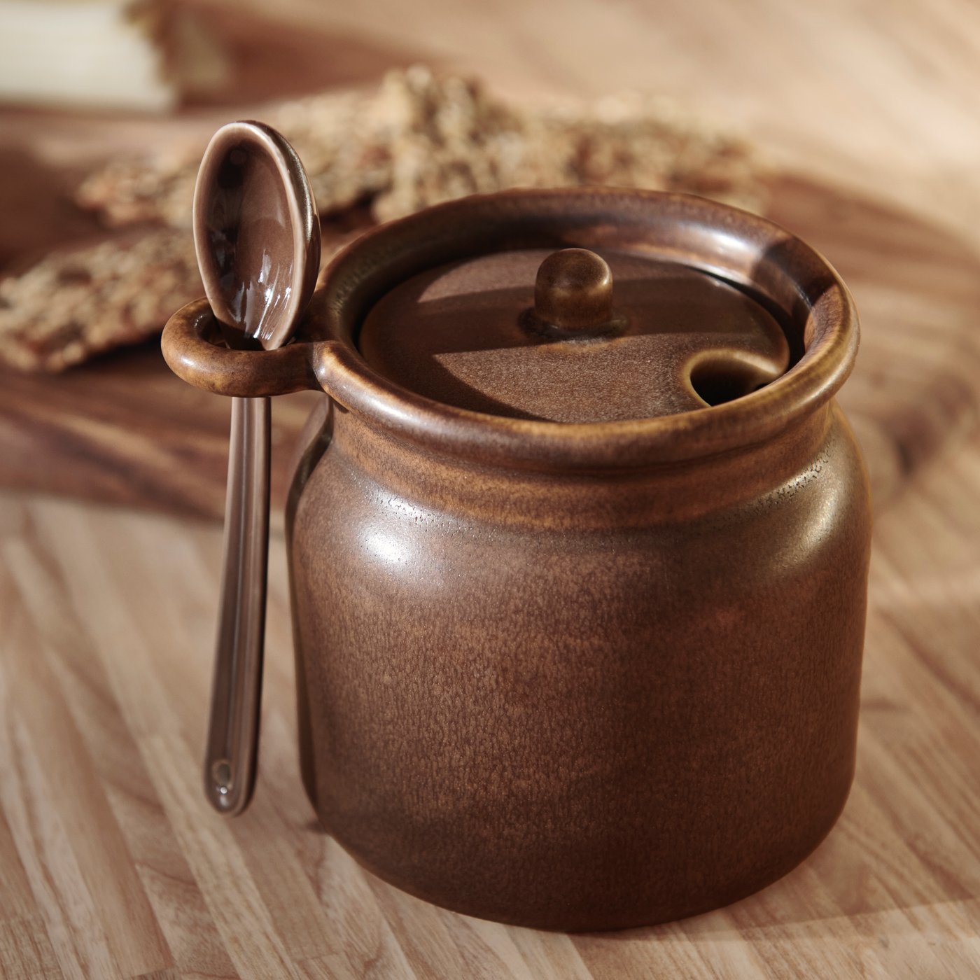 Een bruine KÖSSEBÄR pot met lepel staat op een houten oppervlak. De lepel is netjes geplaatst in de lus aan de zijkant van de pot.