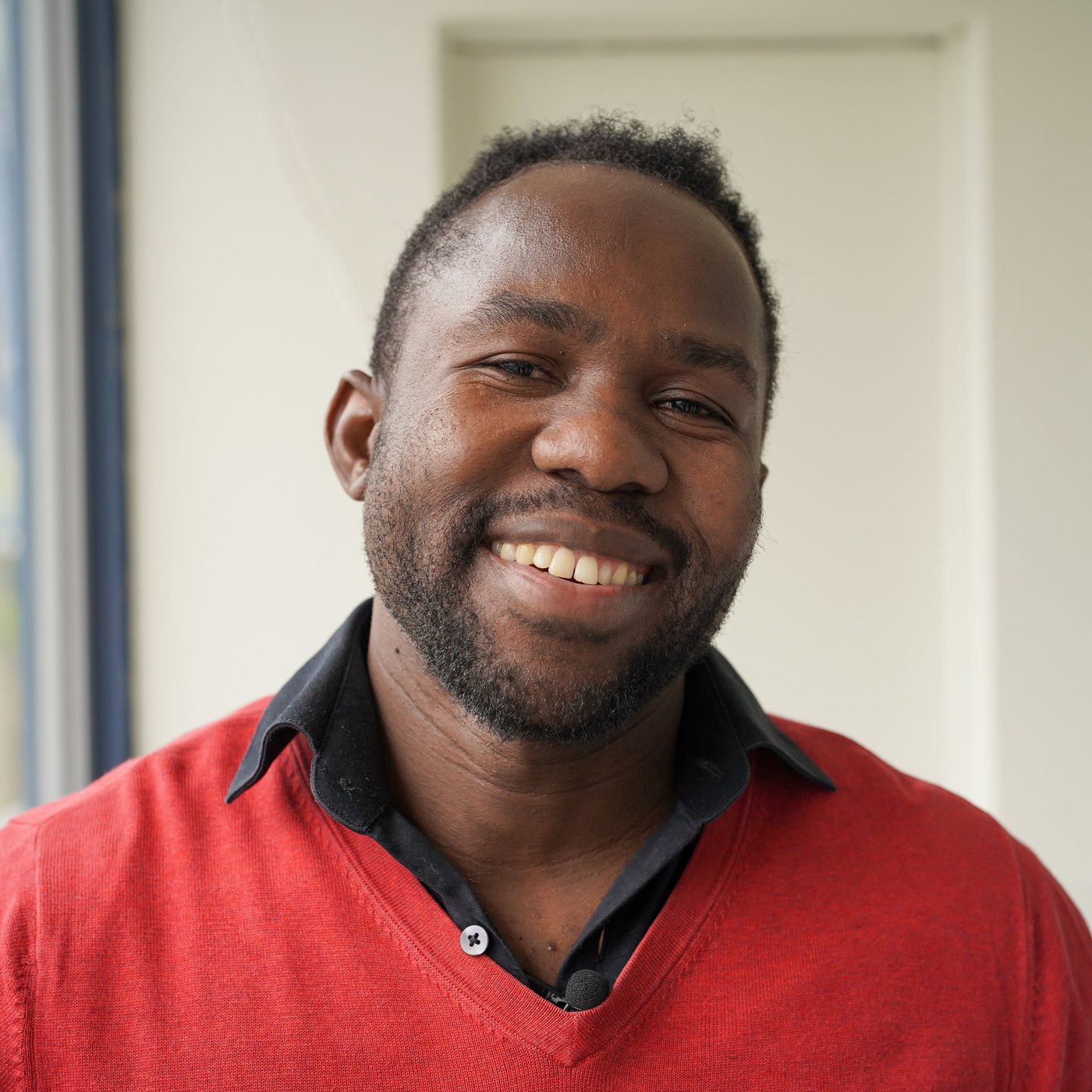 A man sits outdoors in his red shirt and smiles at the camera.
