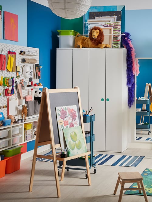 A children's room with two side-by-side white GODISHUS wardrobes and an easel by a pegboard wall of arts and crafts supplies.