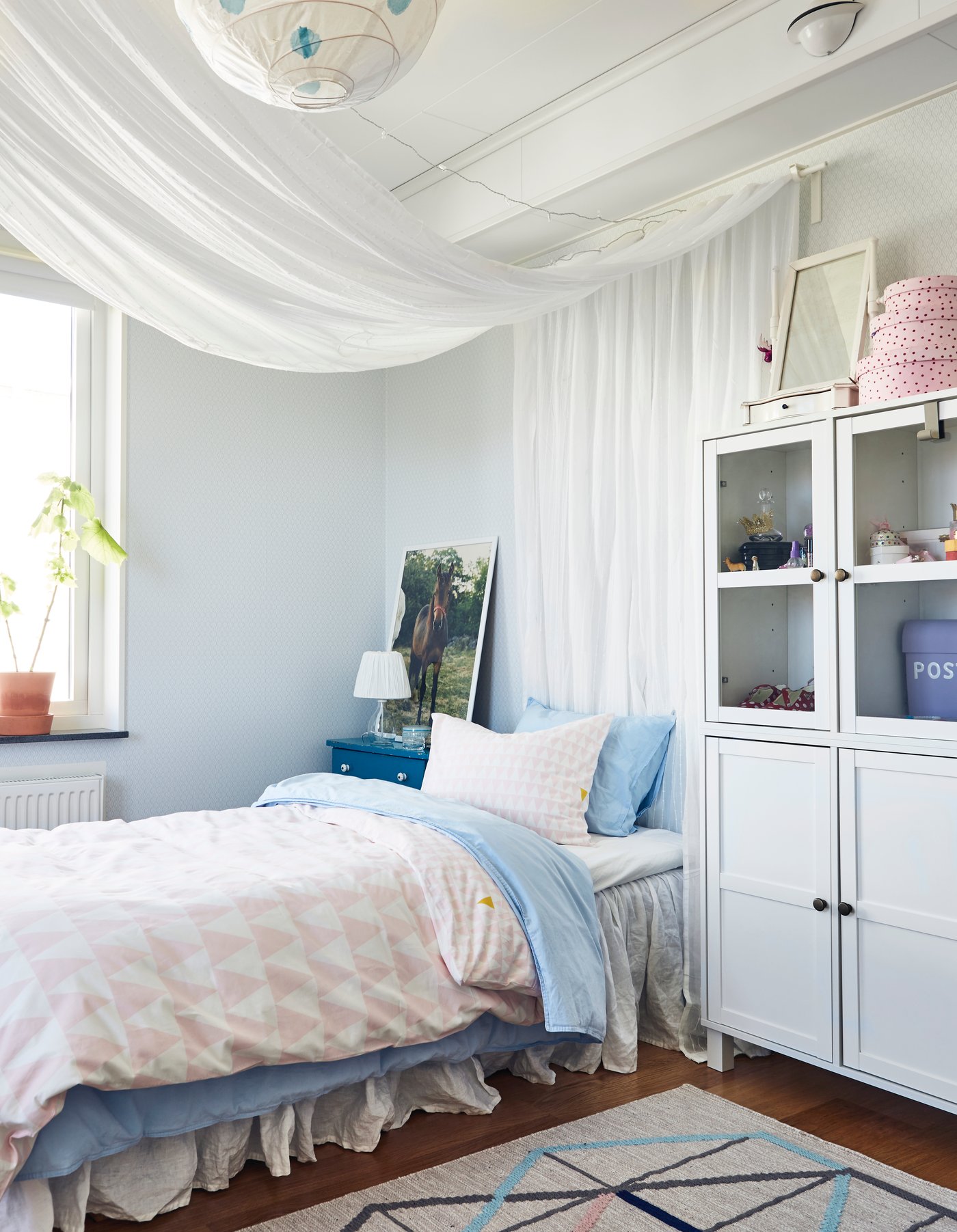 Morbido copripiumino con federa coordinata, caratterizzati da triangoli rosa, piccoli su un lato e grandi sull’altro in cameratta per bambini dai colori pastello. Nella stanza c'è una vetrina bianca, un tappeto, un paralume e una tenda.