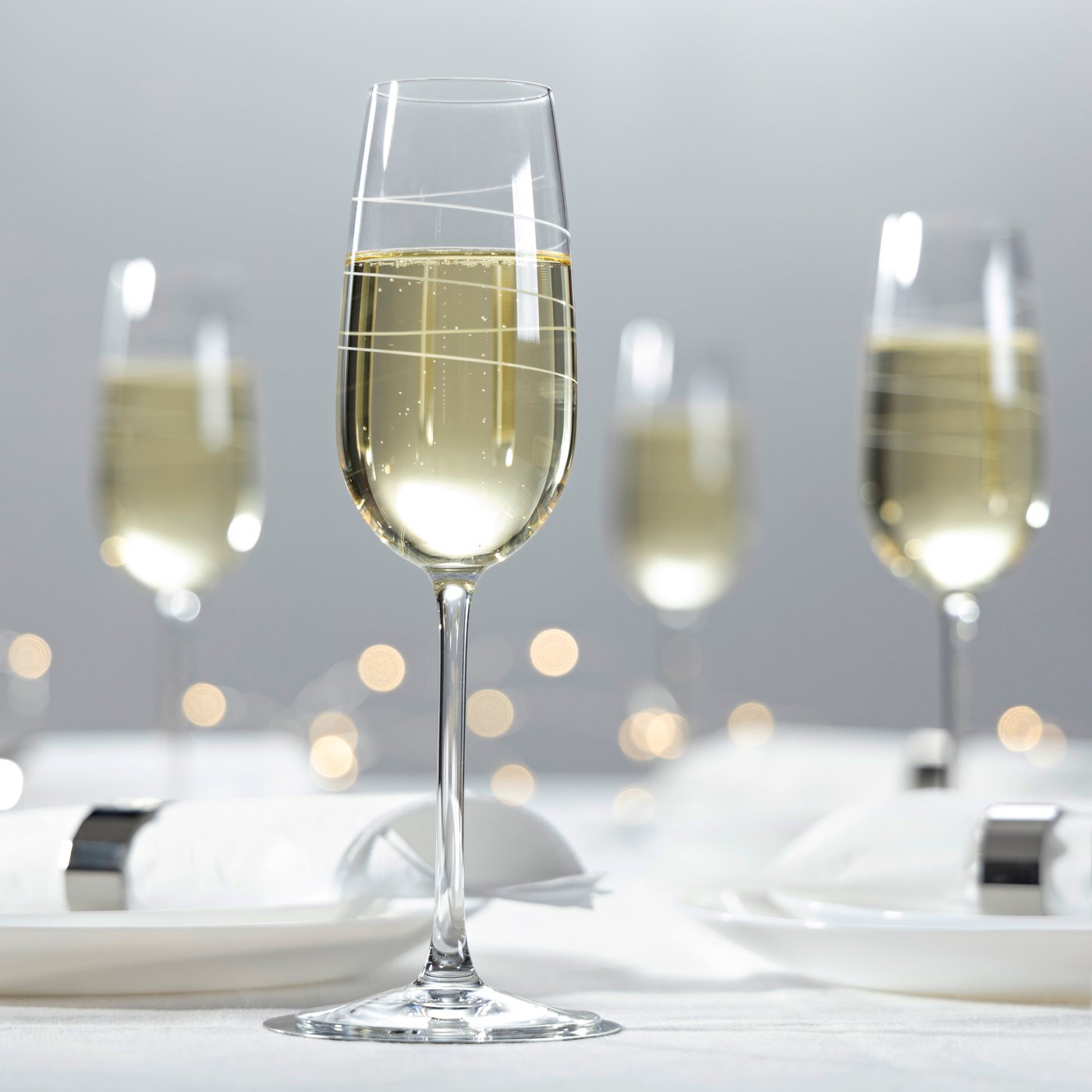 Una tavola elegante sui toni del bianco e dell'argento con luci decorative accese. Ci sono calici di vino bianco pieni.