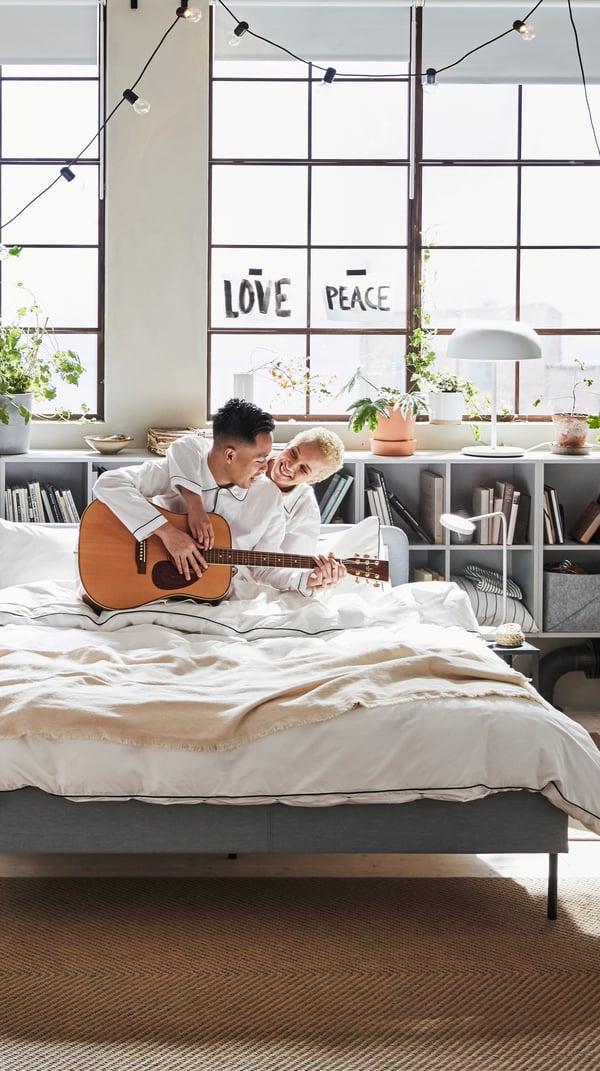Twee personen zitten samen op een bed en hebben een gitaar vast en lachen.