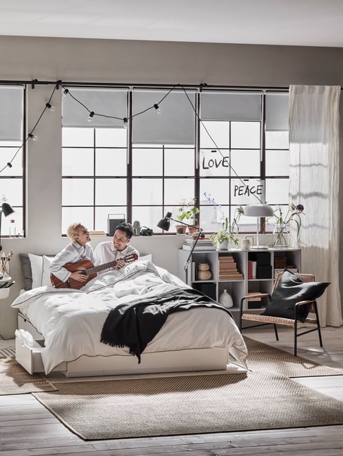 A man and a woman holding a guitar are in a bed with storage, with a rug on the floor, and a table lamp on two cabinets.