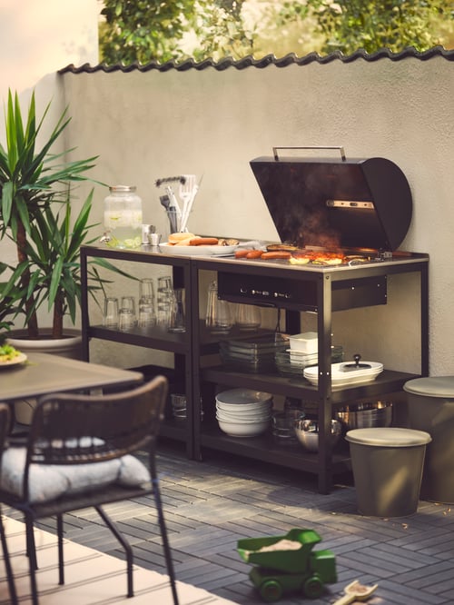 A GRILLSKÄR outdoor kitchen with charcoal barbecue against a wall on a sunny garden terrace. Food is cooking on the grill.
