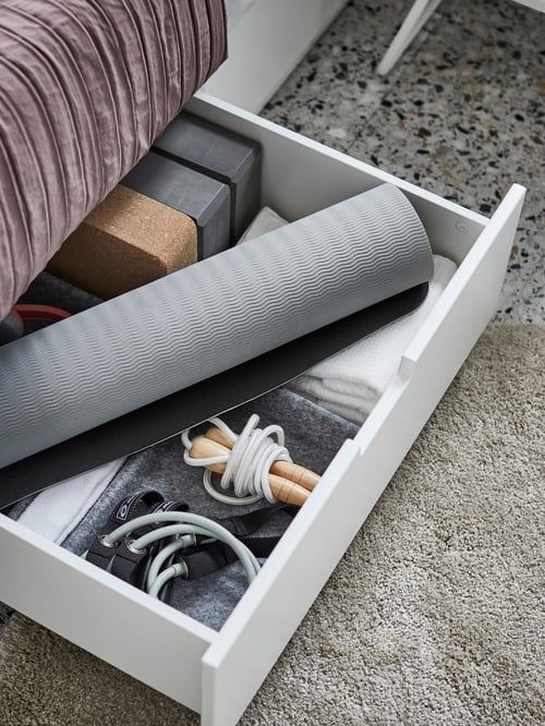 Tiroir ouvert avec un tapis de yoga et d’autres matériels de sport dans un cadre de lit NORDLI avec rangement, sur un tapis gris.
