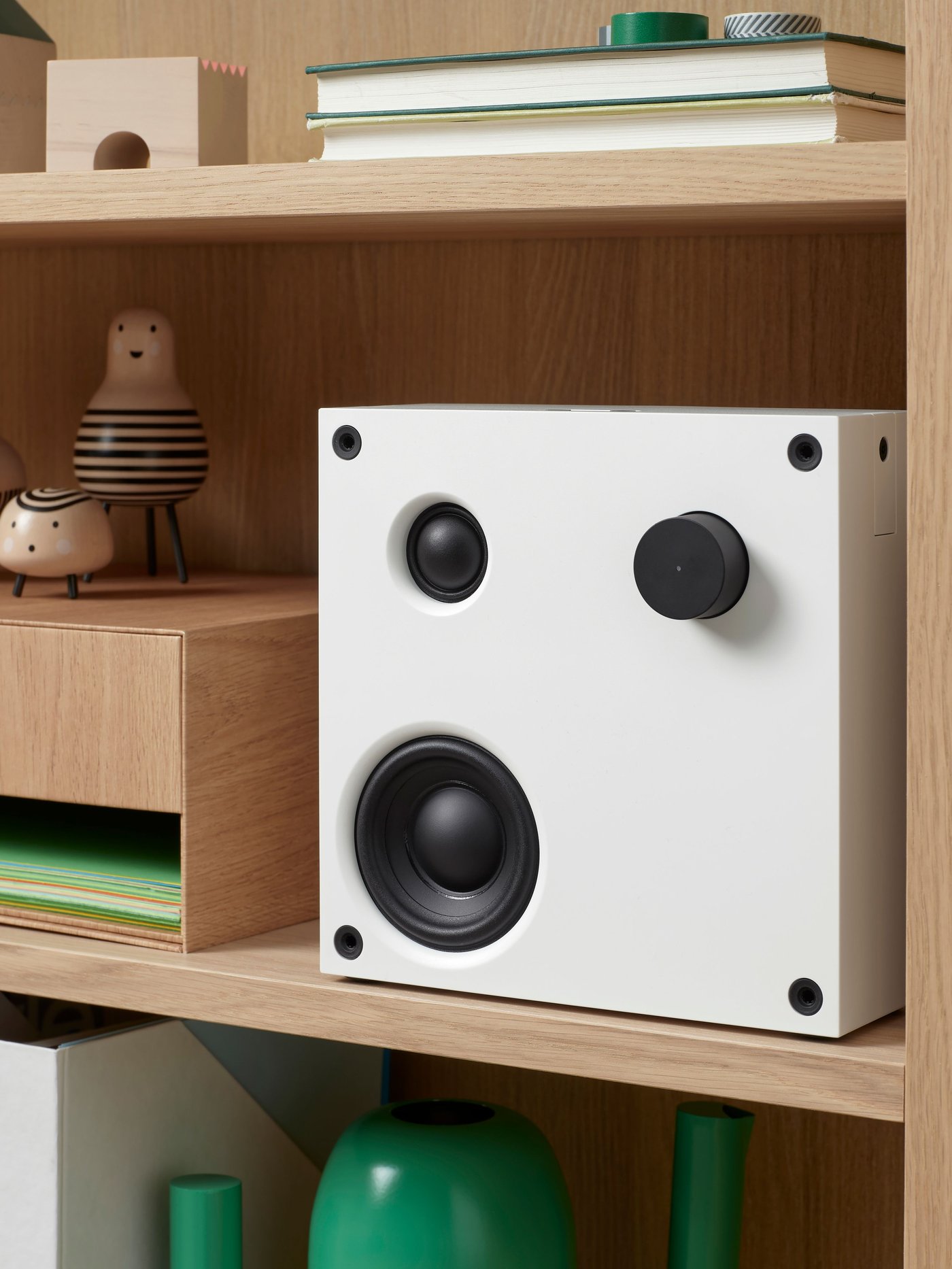 A square, white VAPPEBY Bluetooth speaker with black details like knobs and screws, is on a wooden shelf by decorative items.
