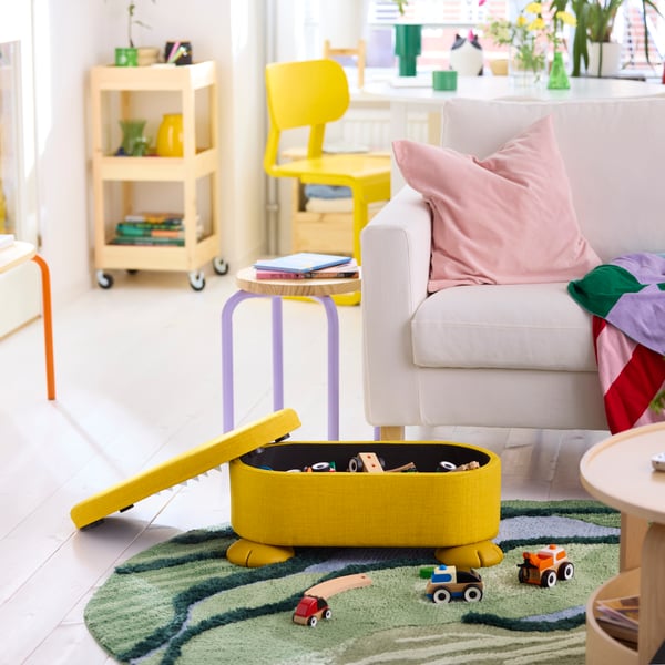 Toys fill a yellow GRESIMOJS pouffe set on a round rug. A white sofa is behind the pouffe. Its lid leans against the pouffe.