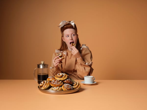 Une personne qui mange une broche à la cannelle avec une tasse de café près d’un plateau de brioches à la cannelle.