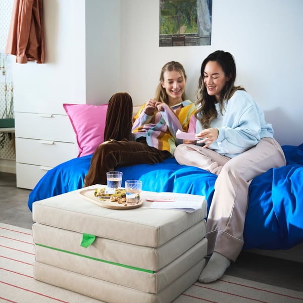 Two students sitting talking in a bed with a SLÄKT pouffe in front of them holding a tray with snacks and water.