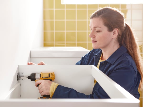 An IKEA co-worker is mounting a commode on to a bathroom wall with a screwdriver.