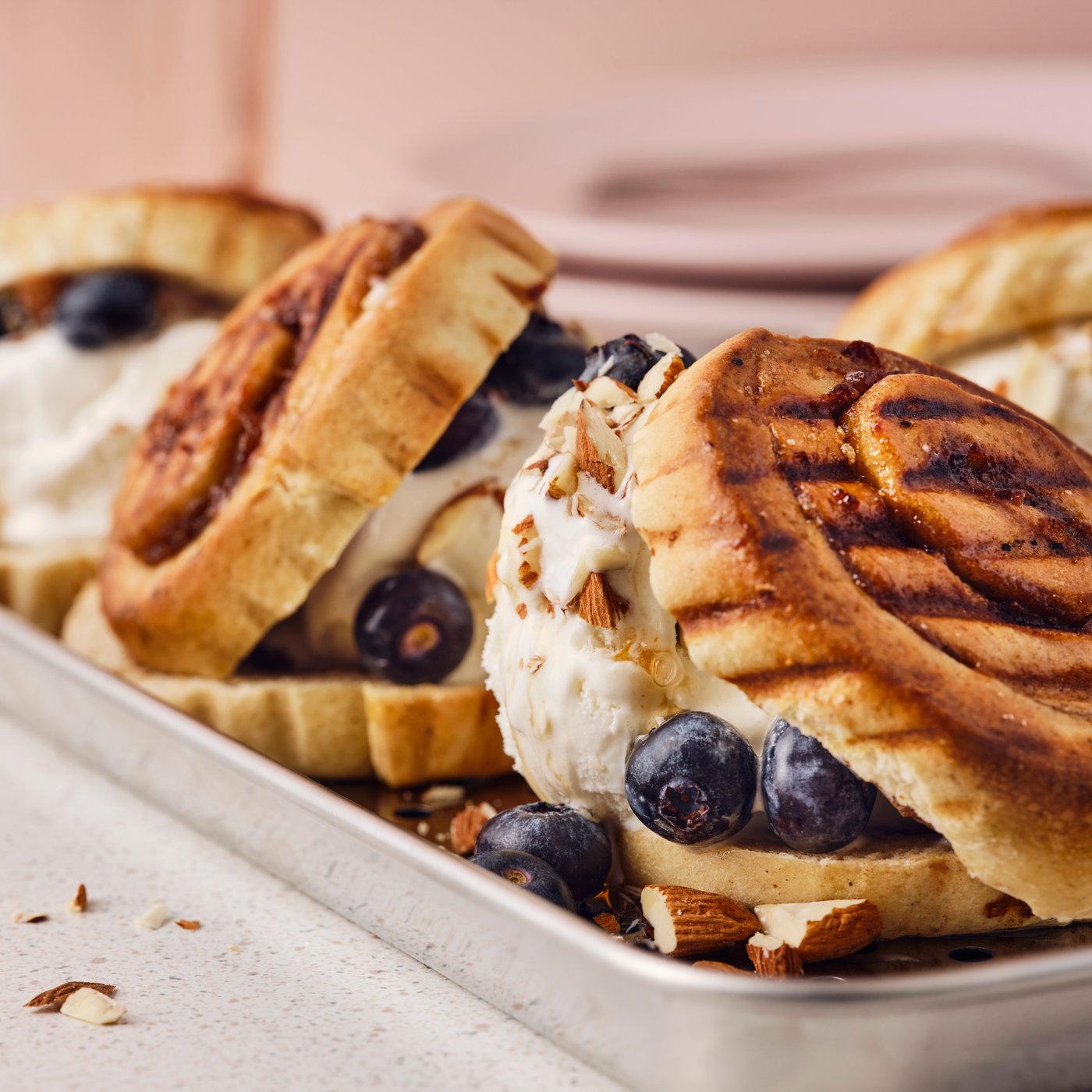 Three grilled KAFFEREP cinnamon buns, ice cream, blueberries, maple syrup and chopped almonds on a metal serving tray.