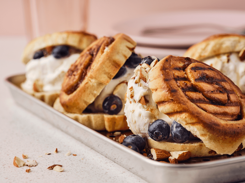 Drei gegrillte KAFFEREP Zimtschnecken, Eis, Heidelbeeren, Ahornsirup und gehackte Mandeln sind auf einem Metalltablett zu sehen.