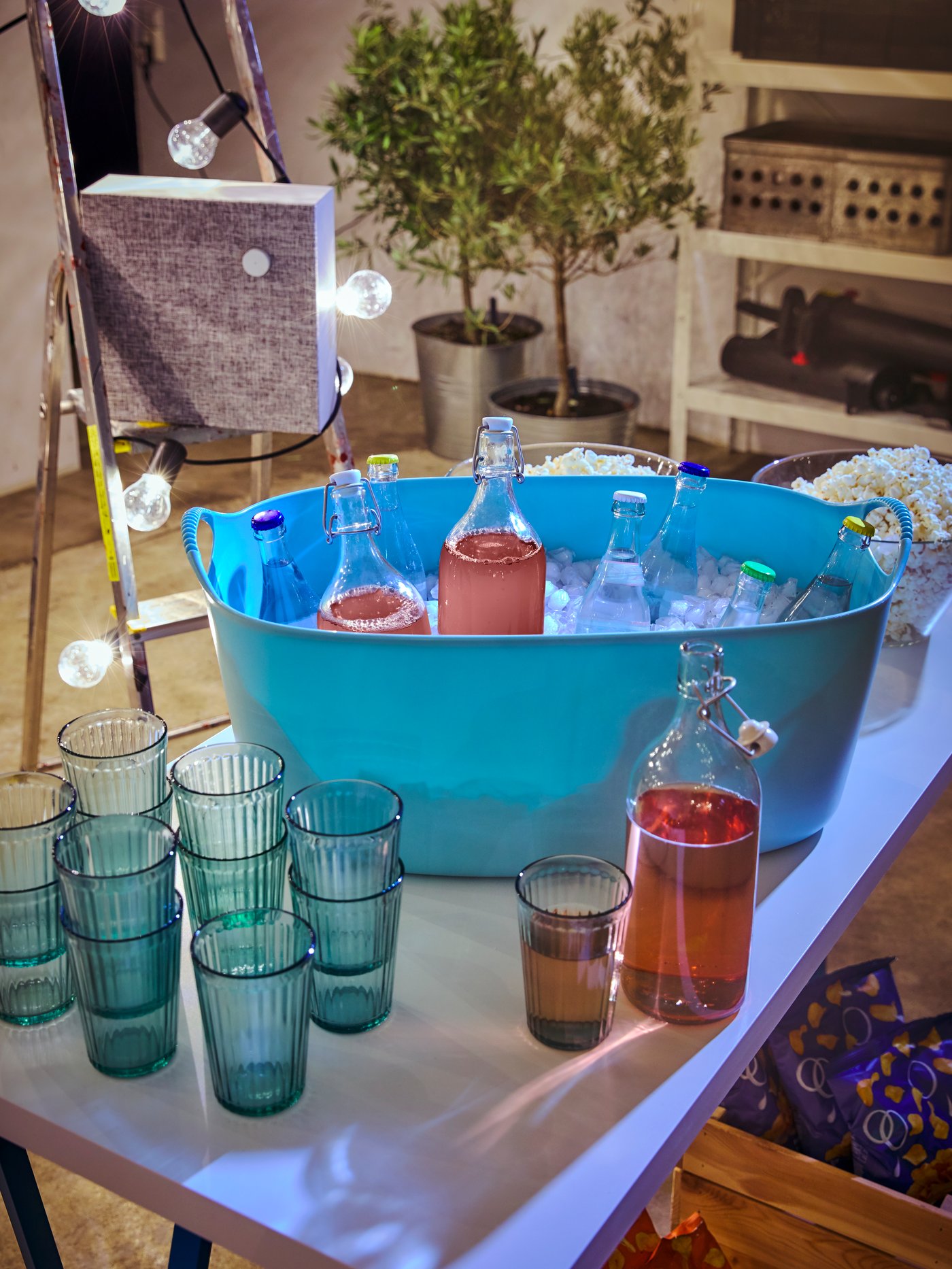 En vasketøjskurv er fyldt med isterninger og karafler fyldt med drinks og ved siden af står blå glas og en højtaler. 