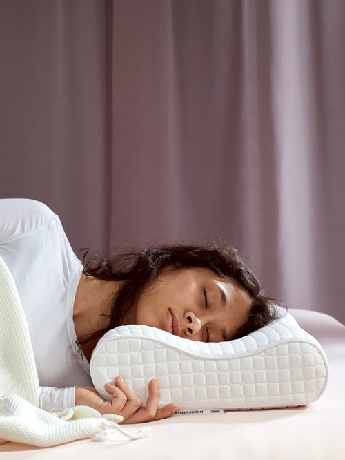 A woman asleep on her side in front of a dark lilac curtain with her head on a ROSENSKÄRM pillow without a cover.