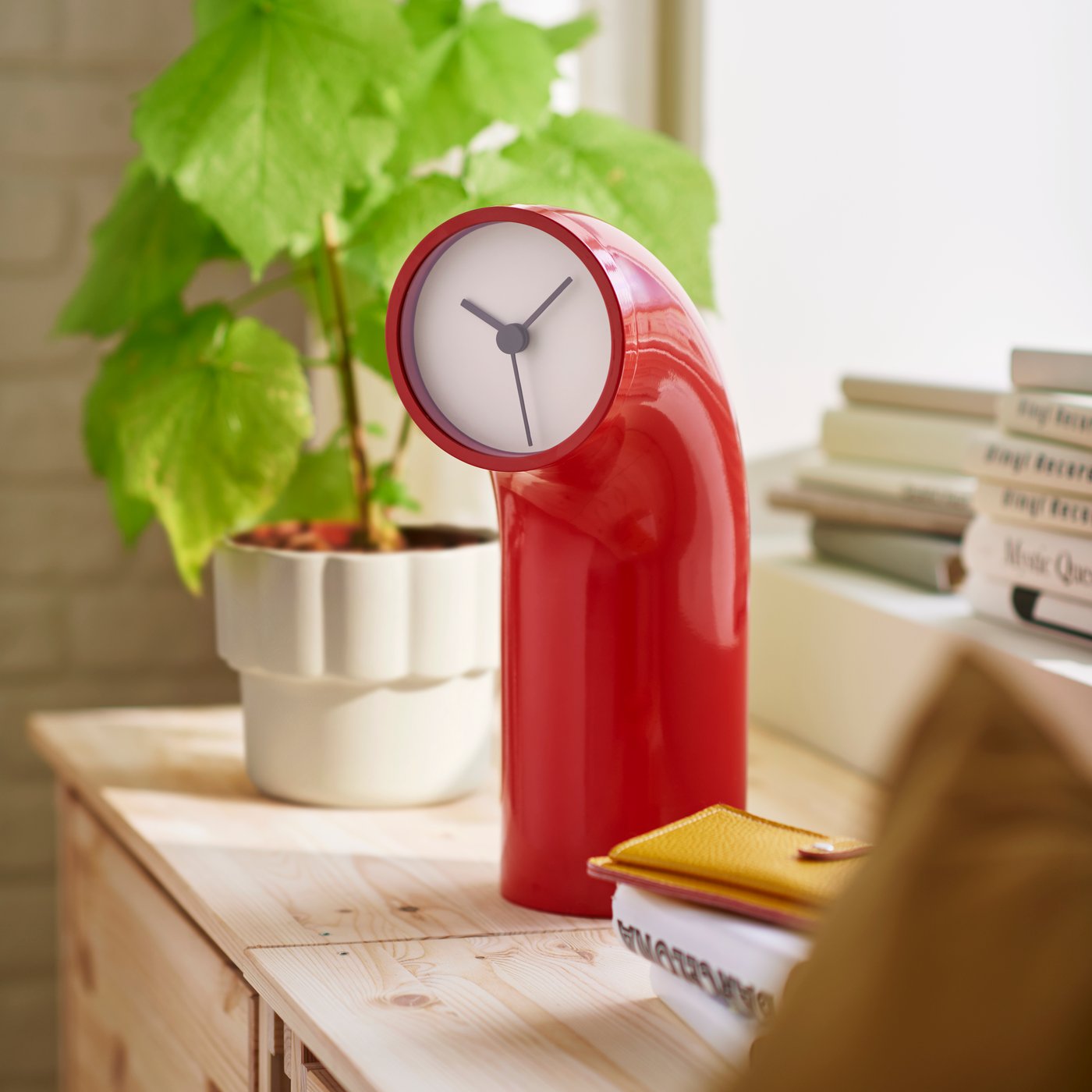 A bright room with a red IKEA PS table clock on a wooden counter beside a plant and stacked books, adding a pop of colour.