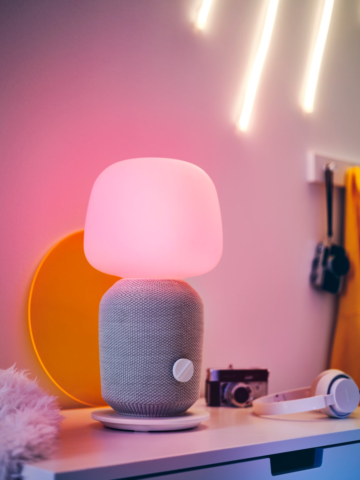 A SYMFONISK table lamp with a built-in WiFi speaker, a yellow tray, headphones and a camera on top of a chest of drawers.
