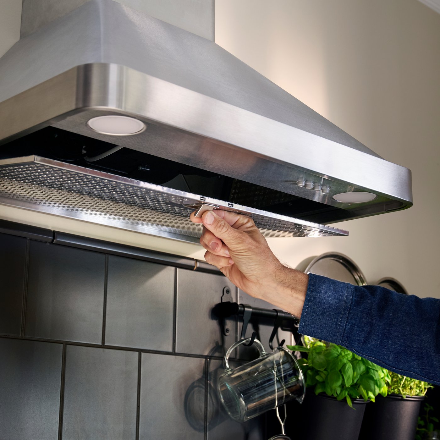 A person in a blue shirt is removing the filter from an extractor hood of stainless steel, mounted in a grey-tiled kitchen.
