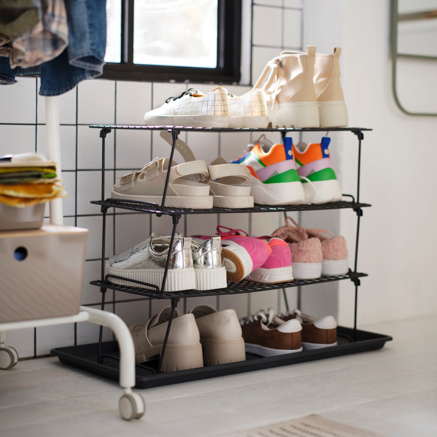 A GREJIG shoe rack on a BAGGMUCK shoe mat is placed by a tiled wall. Several pairs of shoes are placed on the rack and mat.