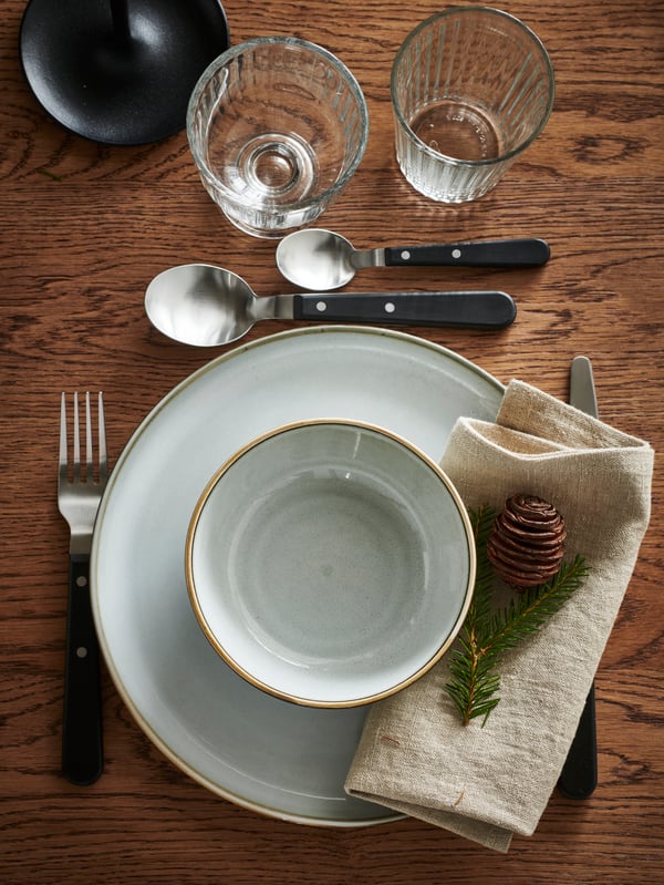 GLADELIG plate setting shown from above on a dark wood table with a folded napkin and pinecone.