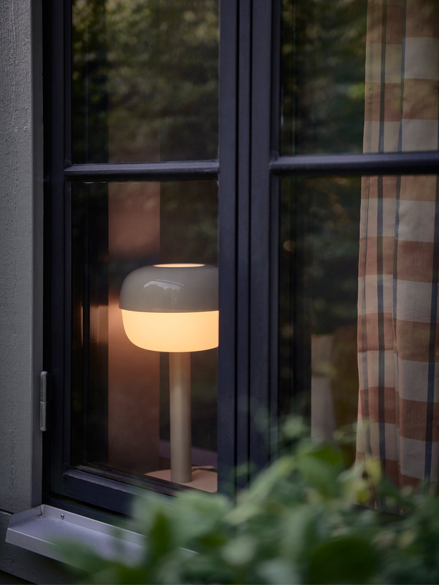 Vy genom ett fönster med svart ram. Inne syns en tänd lampa med vit skärm, randig gardin och grönska utanför.