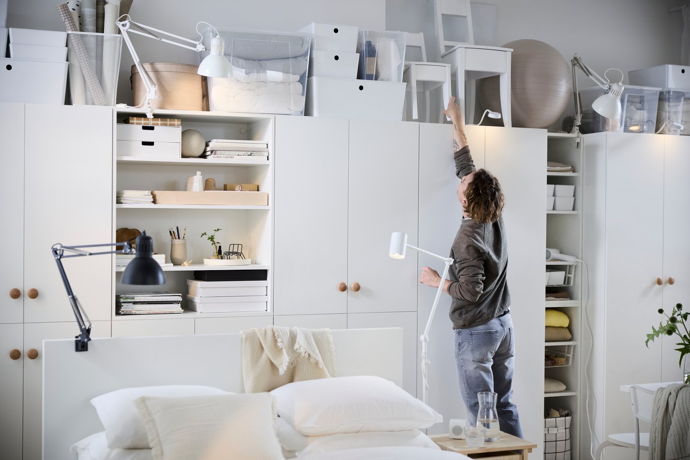 Une personne attrape un objet sur l'étagère supérieure d'un système de rangement modulaire IKEA bien organisé, probablement de la série PAX ou PLATSA, comprenant des armoires blanches avec des poignées en bois et plusieurs lampes de travail TERTIAL réglables, le tout contribuant à une chambre lumineuse et fonctionnelle.