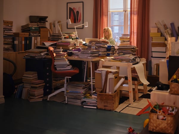 Une femme derrière son bureau en désordre où documents et livres sont empilés sur le bureau, la chaise et les meubles qui l'entourent.