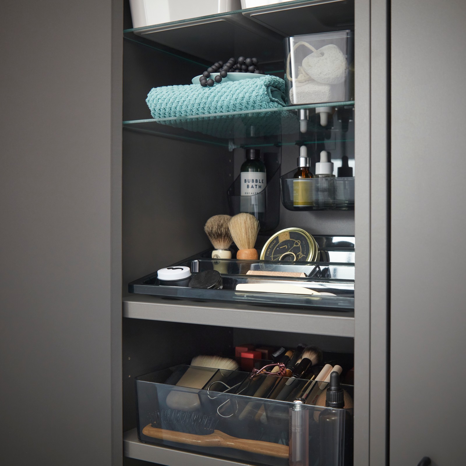 Close-up of a grey HAVBÄCK wall cabinet in a bathroom with a VISSLAÅN grey hanging tray holding organised toiletries.