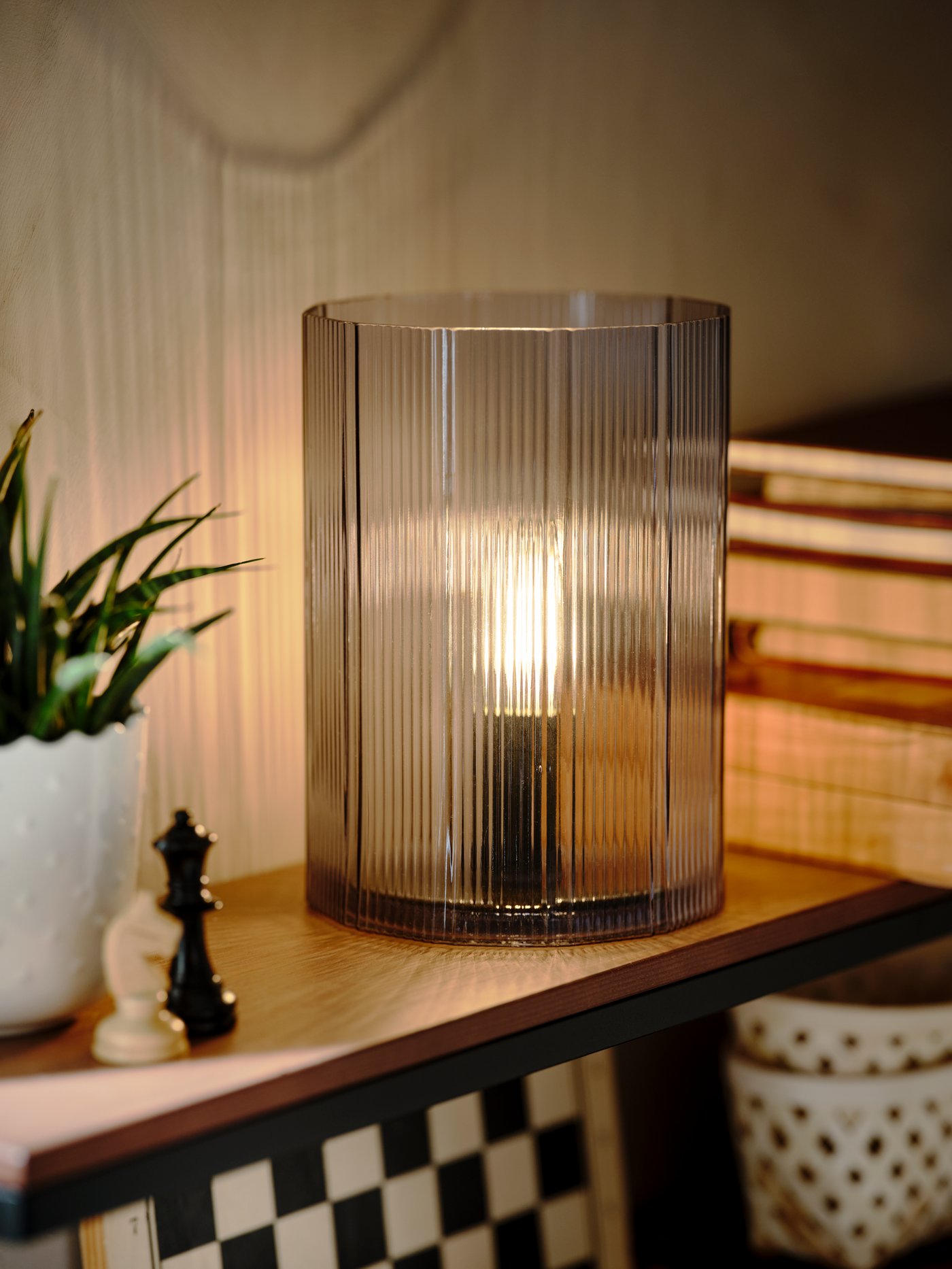 Meja lampu MIKROKLIN kaca berwarna biru menyala di samping buku-buku, pasu tumbuhan dan kepingan catur pada para-para lekap dinding.