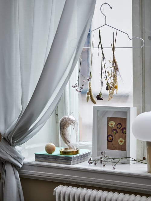 An einem STAJLIG Kleiderbügel, der an einem Fenster hängt, trocknen Blumen. Ein VÄSTANHED Rahmen und eine BEGÅVNING Glasglocke stehen auf dem Fensterbrett davor.